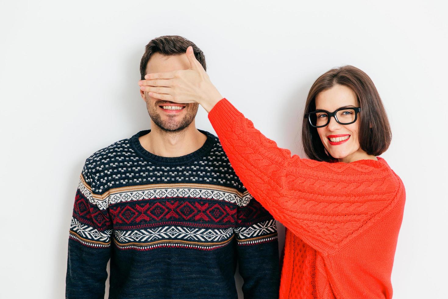 Indoor shot of attractive brunette female with positive expression, covers mans eyes, wear winter sweaters, stand together against white background, have fun. Thats surprise for you, dont look photo