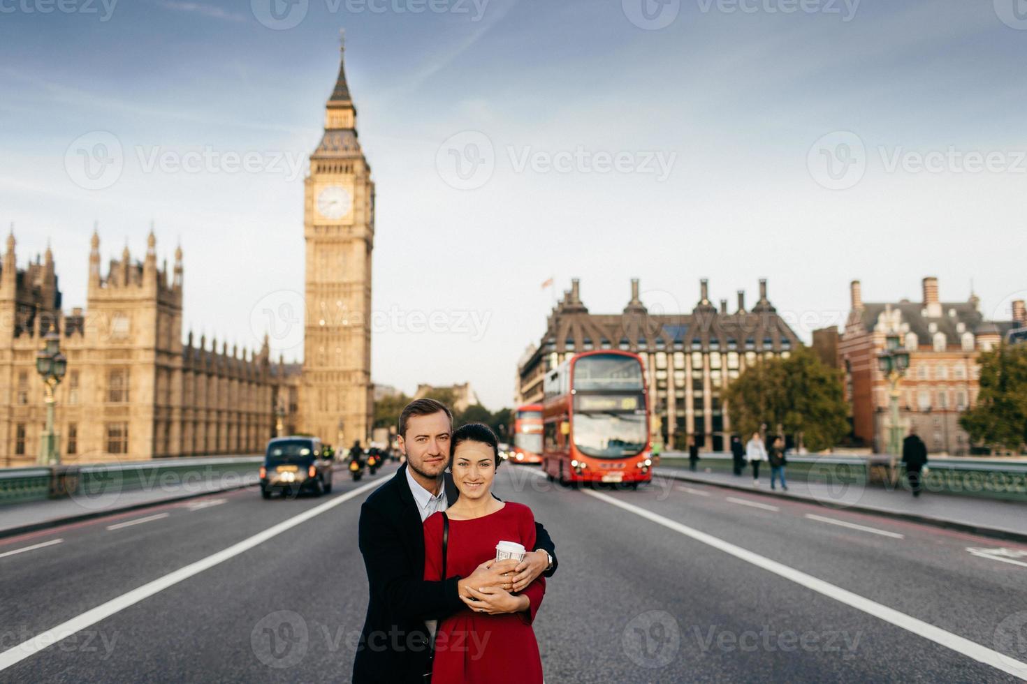 Lovely married couple embrace each other stand on road with Big Ben and London bus background, spend vacations together in Great Britain, recieve pleasant emotions and good impressions photo