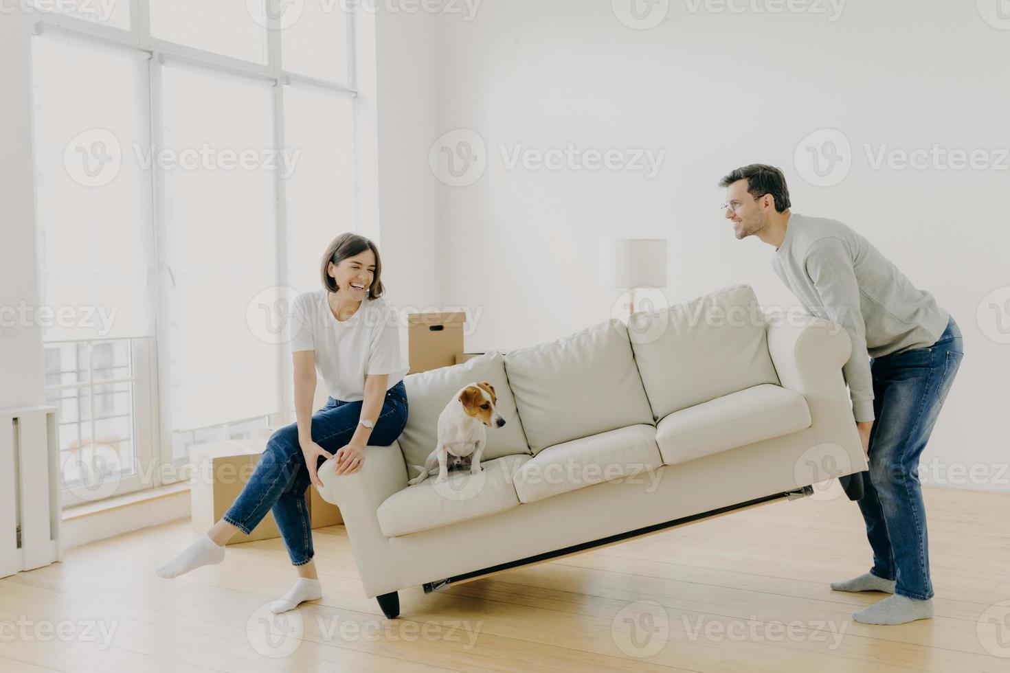 Happy husband and wife place sofa in living room, furnish their first home, help each other in renovation, little dog sits on couch, carry furniture together. Remodeling and renovation concept photo