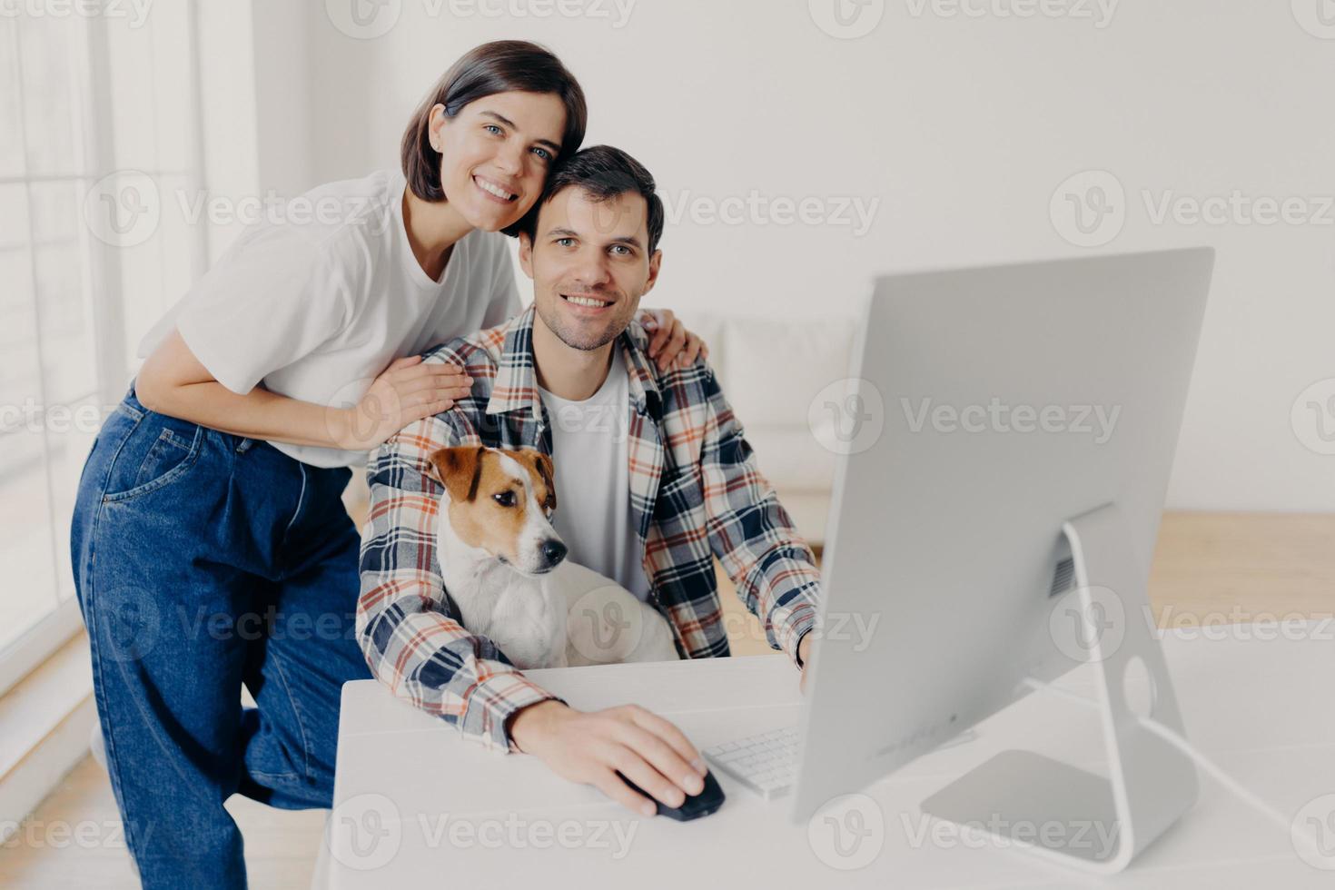 Indoor shot of happy family couple pose near computer monitor, work together remotely, have own business, earn money online, look gladfully at camera, play with favourite pet. People, technology photo