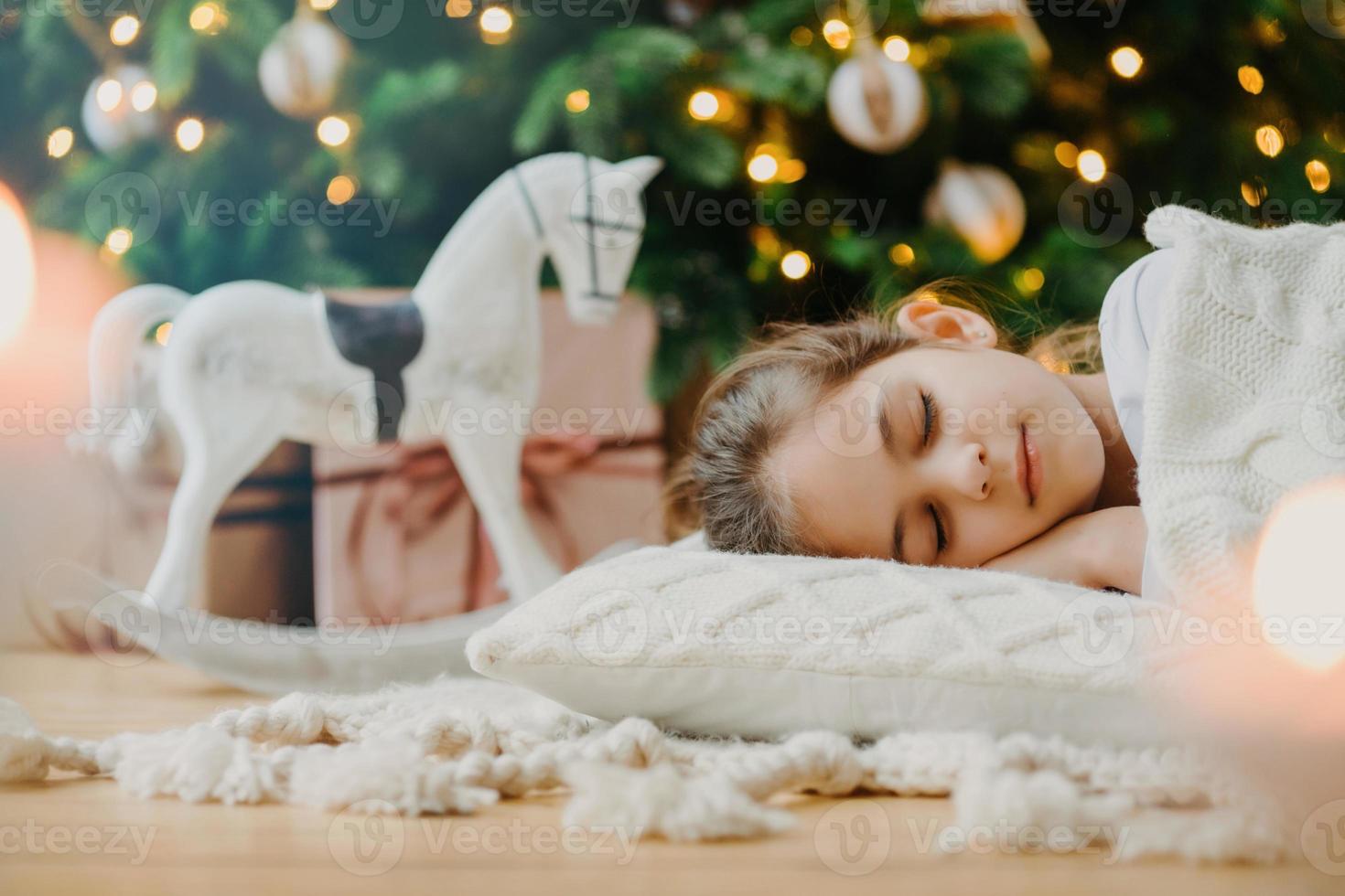 Horizontal shot of relaxed girl covered with white sweater, has pleasant dreams, lies under decorated Christmas tree, toy rocker and gift boxes around. Kid at home. Cozy holiday. Domestic atmosphere photo