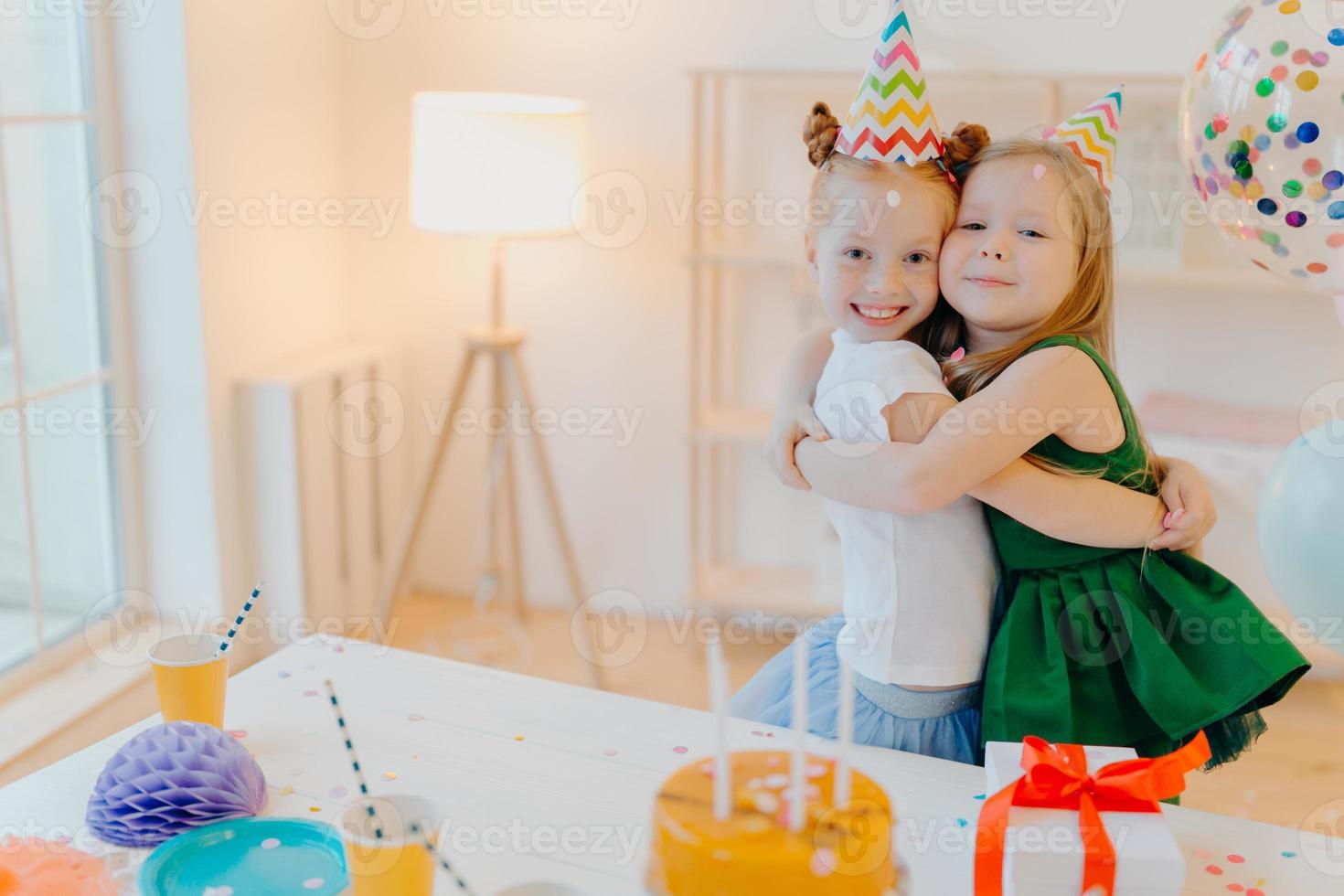 Friendly two girls embrace and have good relationship, stand near festive table with cake, celebrate birthday together, stand in living room. Glad female sisters enjoy holiday, special occasion photo