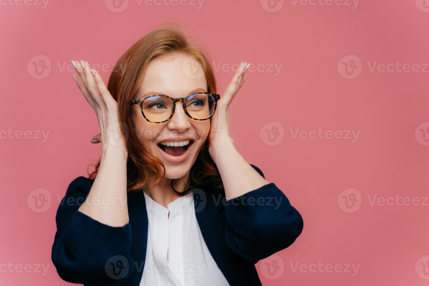 mujer pelirroja sonriente positiva cubre los oídos, feliz de escuchar algo ruidoso y divertido, usa anteojos transparentes, ropa elegante en blanco y negro, se para sobre fondo rosa con espacio libre para texto foto