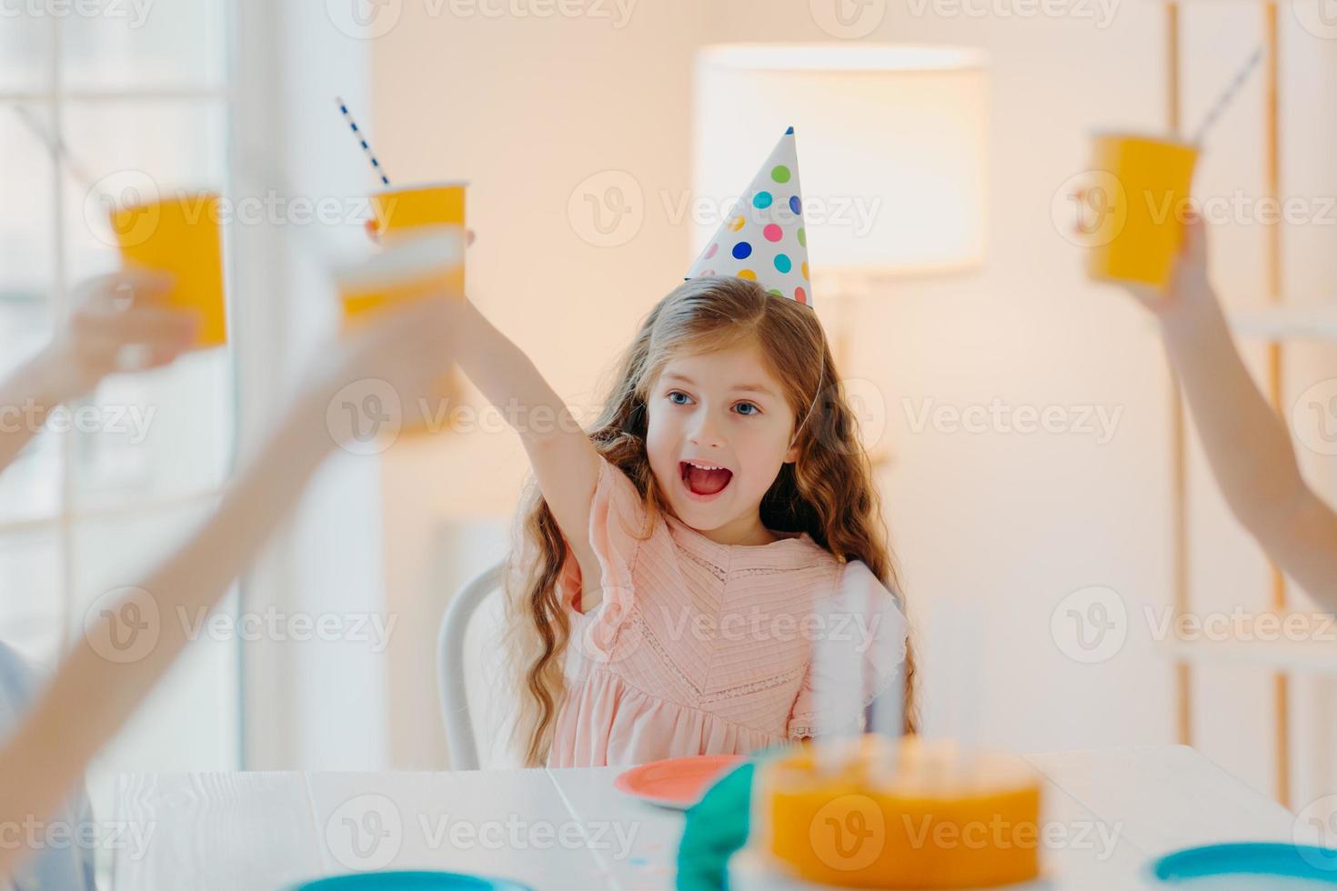 Happy ginger girl wears party hats, clink cups with drink, has fun with friends, celebrate birthday together, pose in decorated room at home. Children, celebration and holiday concept photo