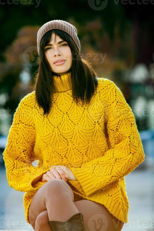 Beautiful European brunette dressed in a yellow woolen sweater and hat outside. The beautiful girl who wears thick stylish autumn clothes in cool weather. photo