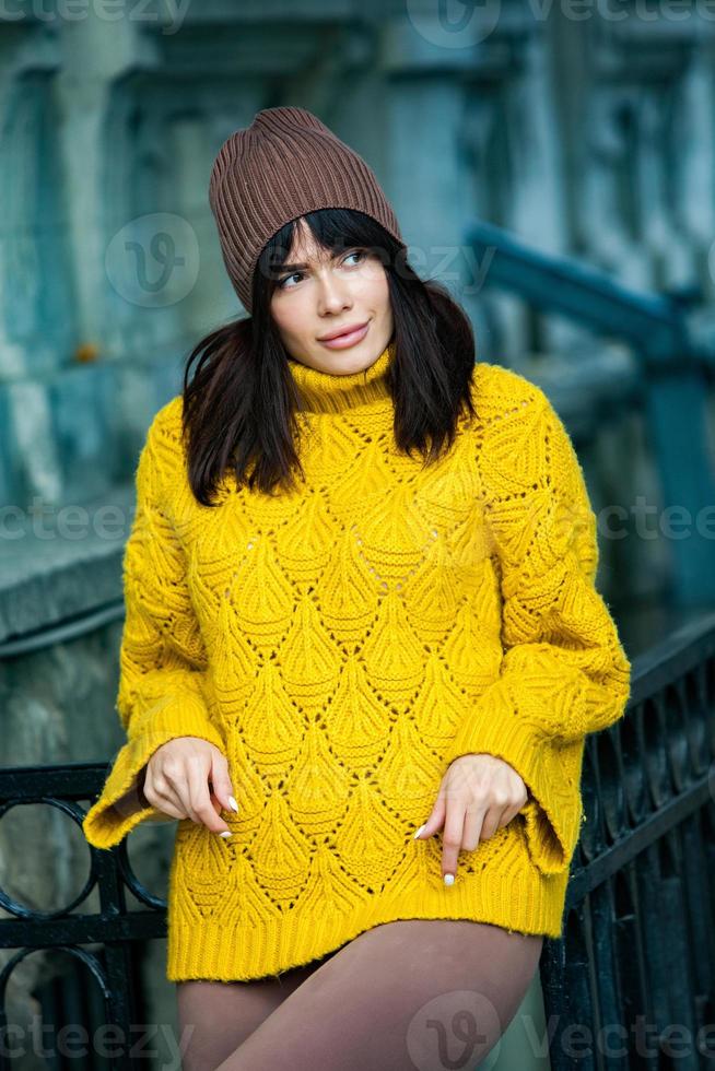 Beautiful European brunette dressed in a yellow woolen sweater and hat outside. The beautiful girl who wears thick stylish autumn clothes in cool weather. photo