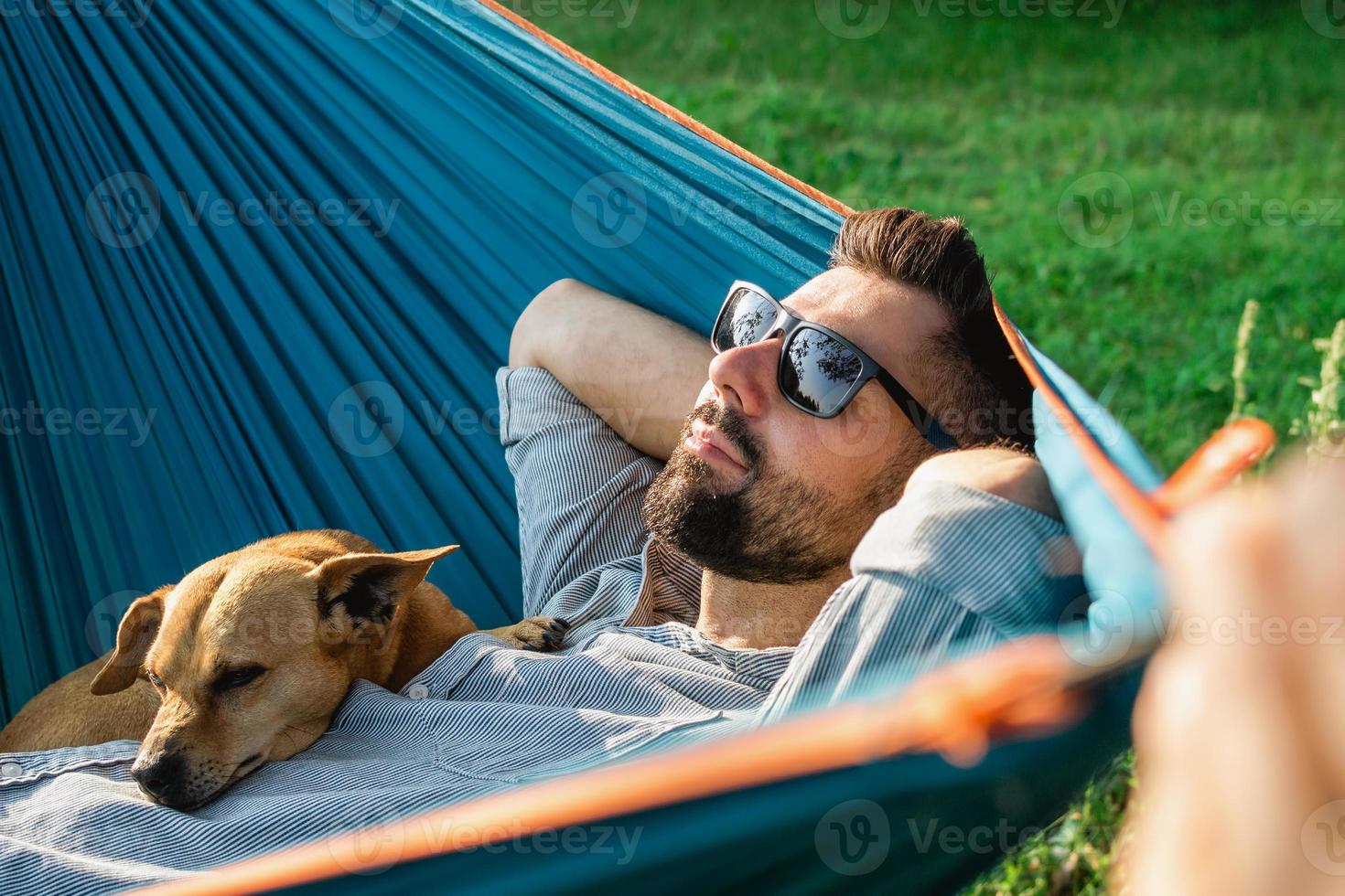 Smiling attractive European man in sunglasses is resting in hammock with his cute little dog. photo