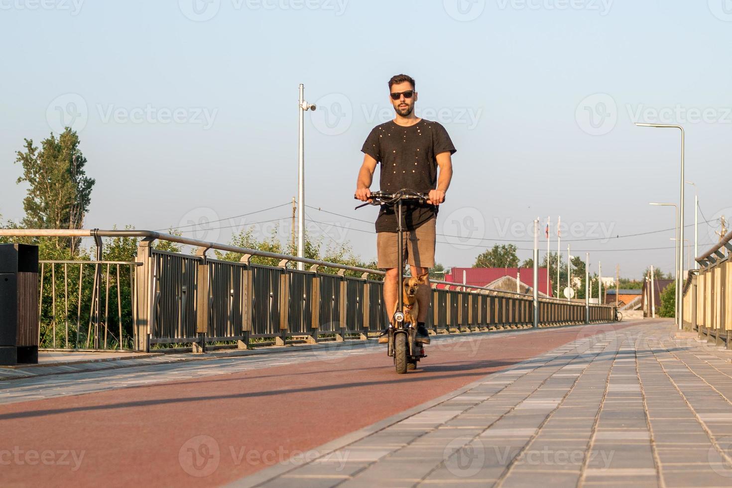 Attractive European man is riding on electric scooter with his small dog. Walking with dog on empty street of city at sunny day. photo