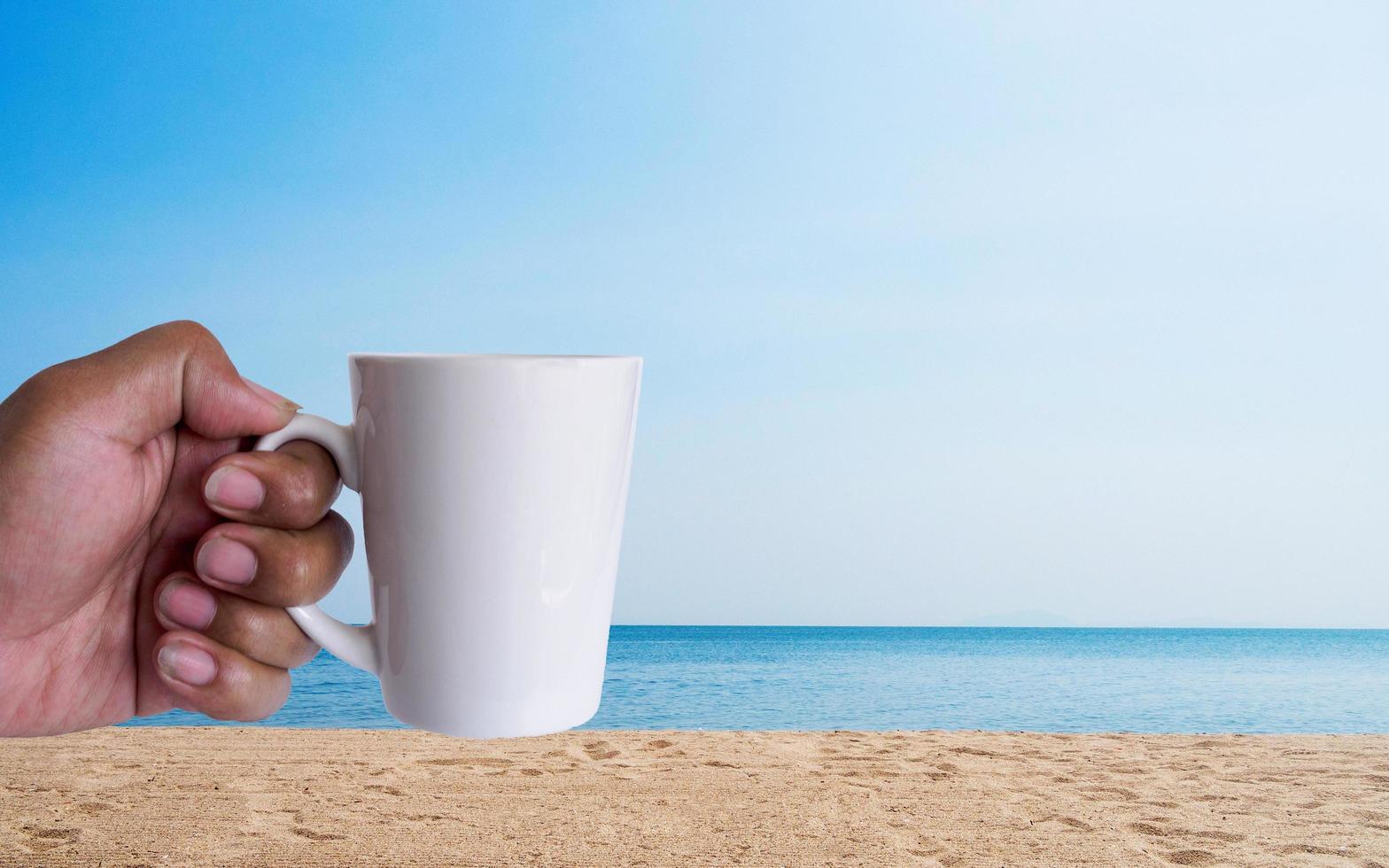 Man hand holding white peper coffee cup takeaway yellow lid, filled with hot black coffee no sugar milk, ready drink, refreshing. aroma awake fresh to work placed enjoy on sea beach blue sky sunshine. photo
