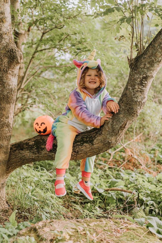 little girl in a rainbow unicorn costume kigurumi is sitting on a tree with pumpkin basket for sweets against the background of a forest. Halloween concept. space for text. High quality photo