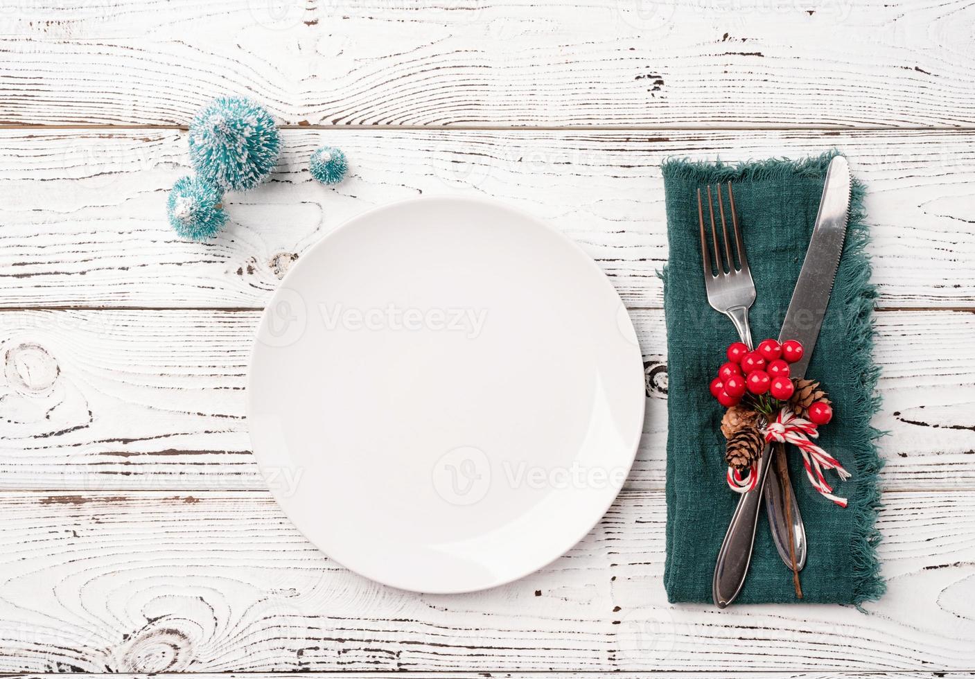 Christmas table setting with white dishware, silverware and red and green decorations on white wooden background. Top view. photo