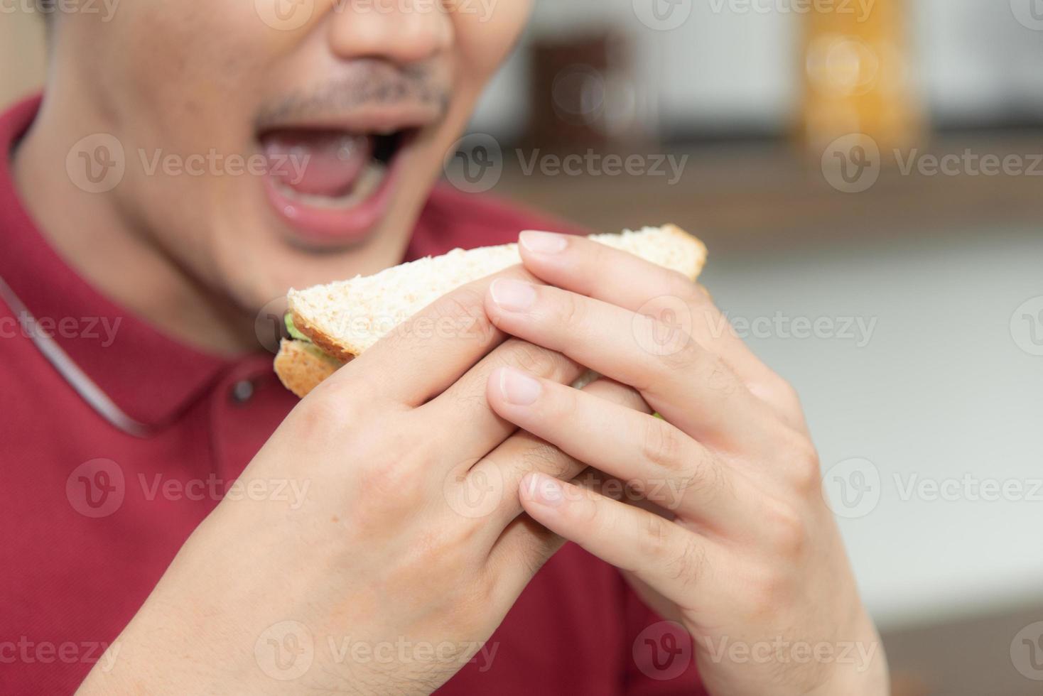 Asian smiling young man with casual  red t-shirt enyoy having breakfast, eating sandwich, Young man cooking food and drink in the loft style kitchen room photo