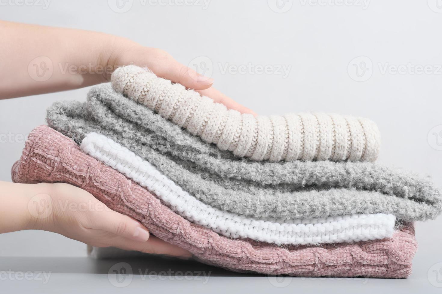 female hands holding a pile pastel color jumpers and other knitted clothing. winter apparel, trendy pale textured knitwear. cozy fall pullovers. photo