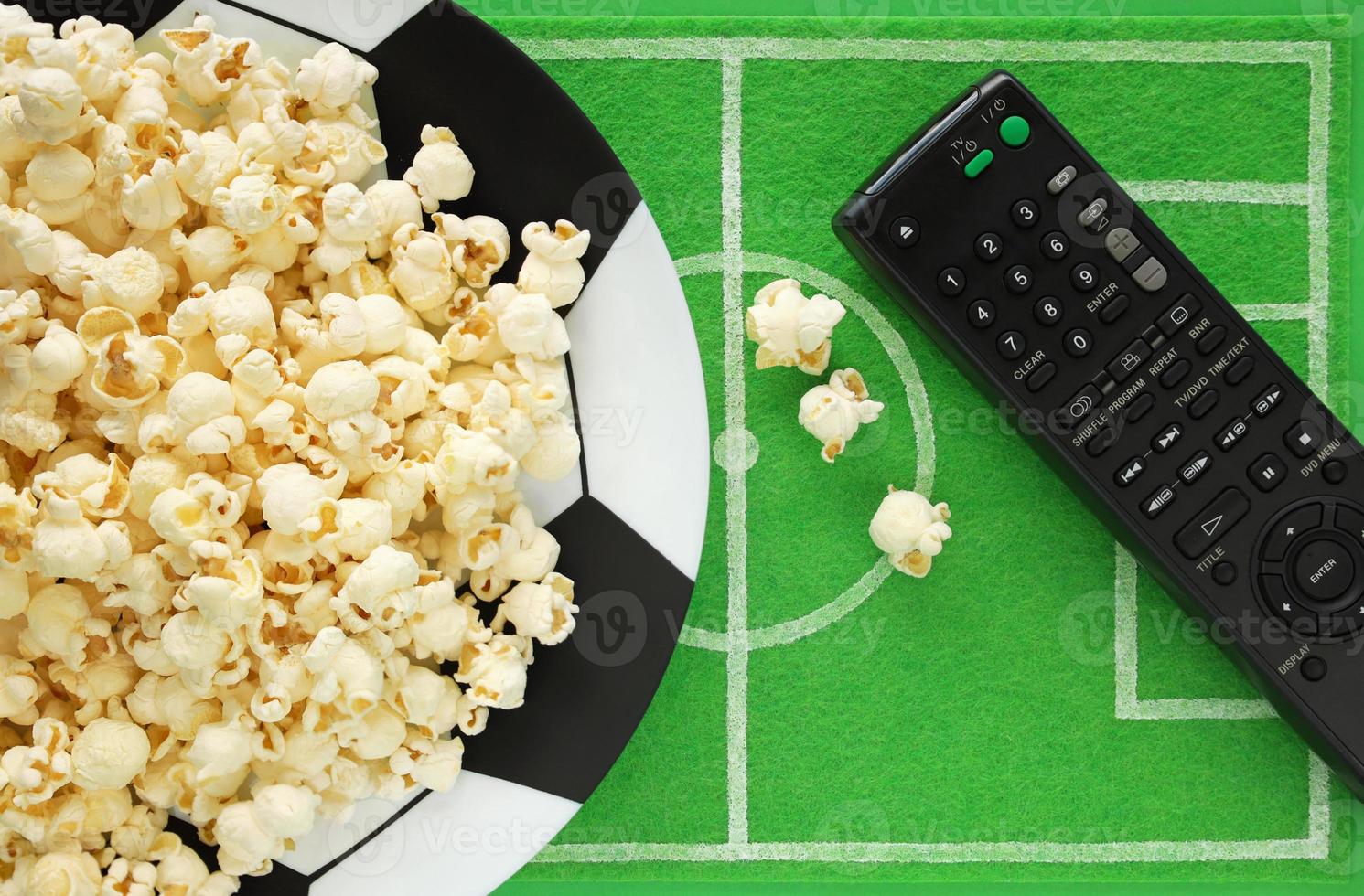 Football soccer party table. Popcorn on big plate painted like soccer ball and dish mat like football field made of green felt, top view. TV remote for watching foorball match broadcast. photo