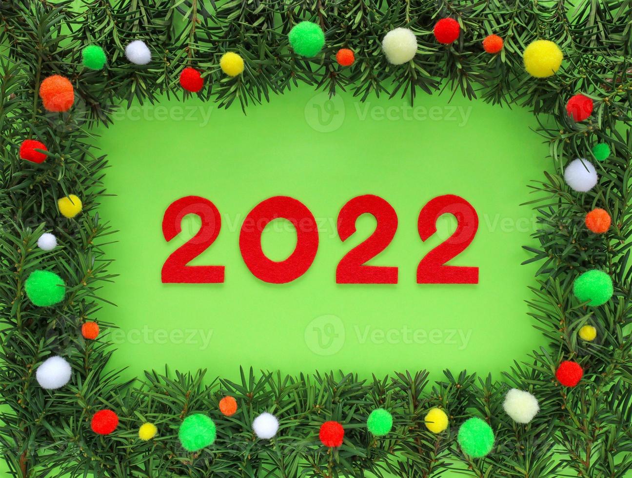 Red felt new year numbers 2022 in frame made fir branches around on light green background. Border decorated colorful pom poms - white, yellow, orange, red, green. Review of the year. Christmas 2022. photo