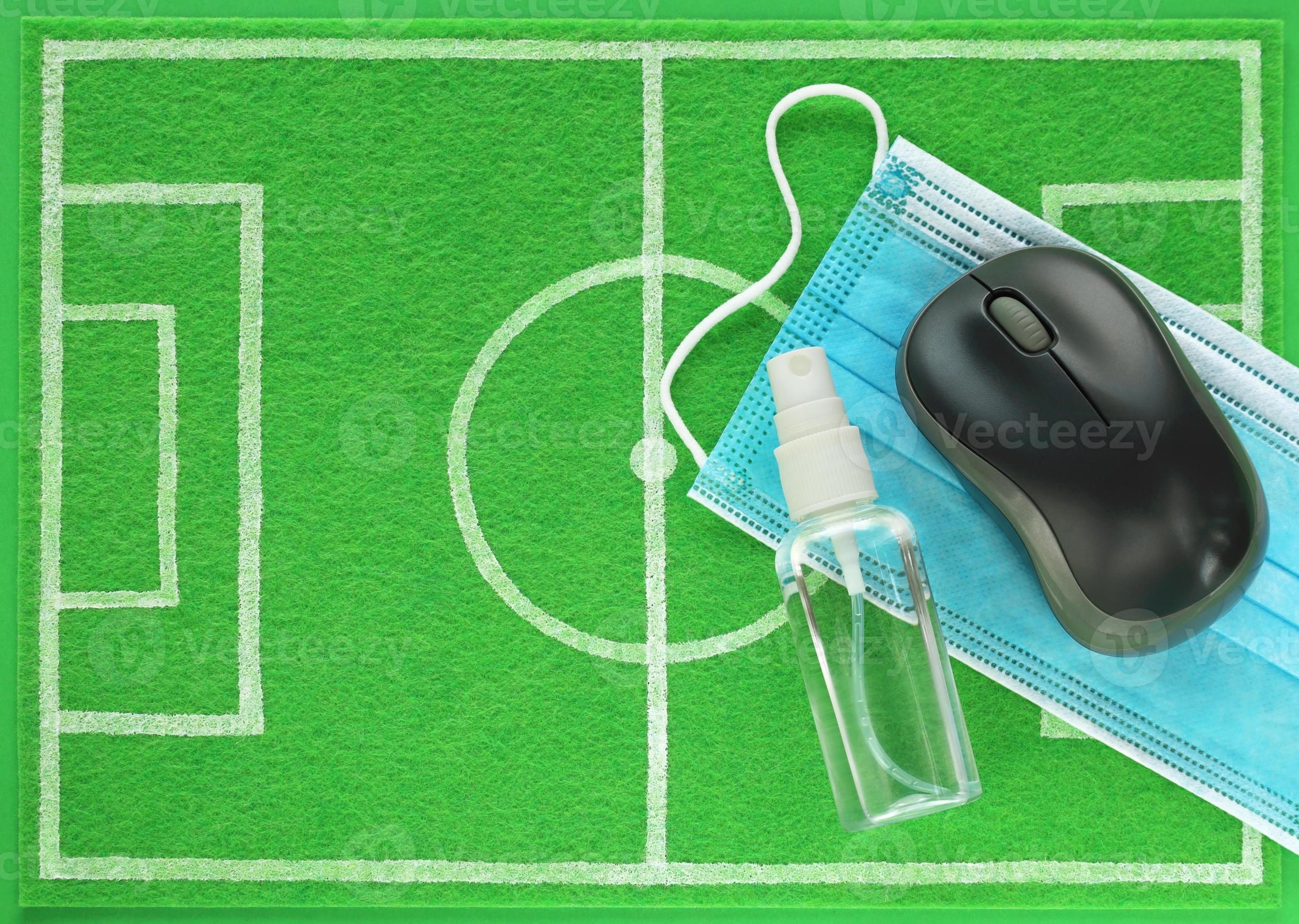 Computer mouse and sanitizer with blue medical mask placed on mini football field made of green felt, top view