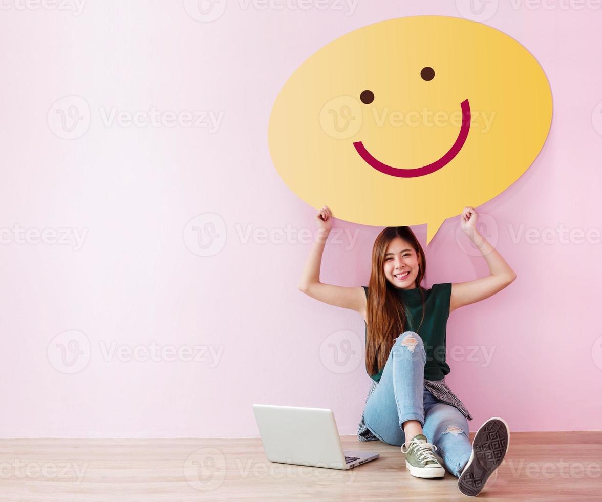 Happy Customer Concept. Review and Feedback her Experience for Satisfaction Survey Online. Young Female in Cheerful Posture, Raise up Speech Bubble with Smiley Face. Sit on the Floor with Laptop photo