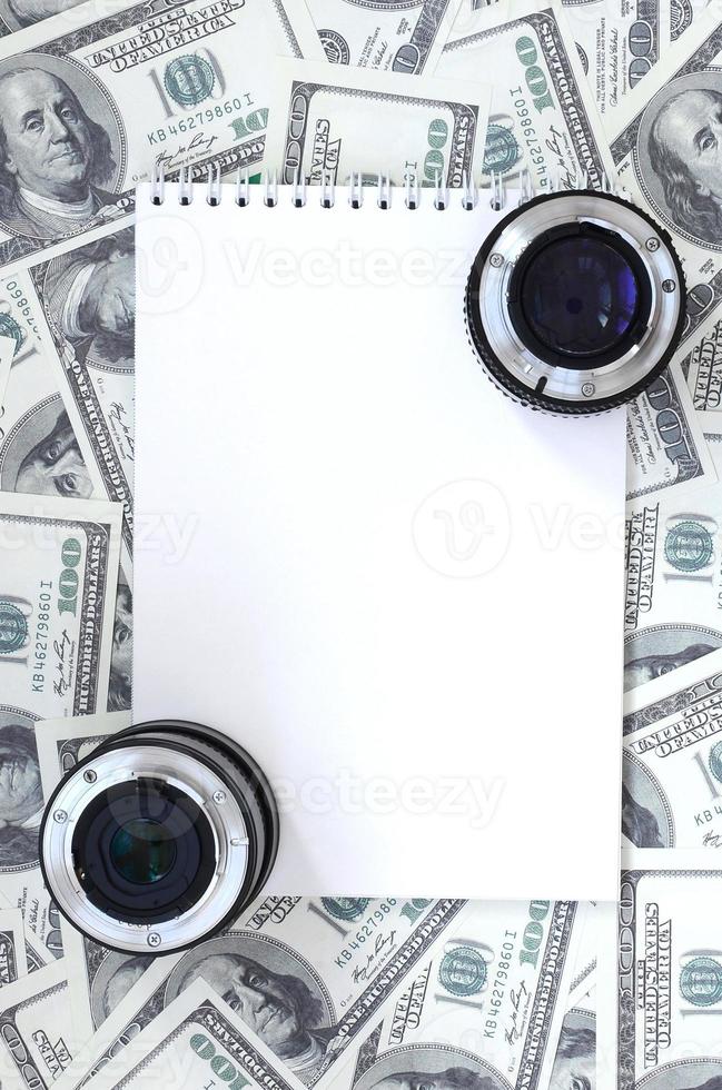 Two photographic lenses and white notebook lie on the background of a lot of dollar bills. Space for text photo