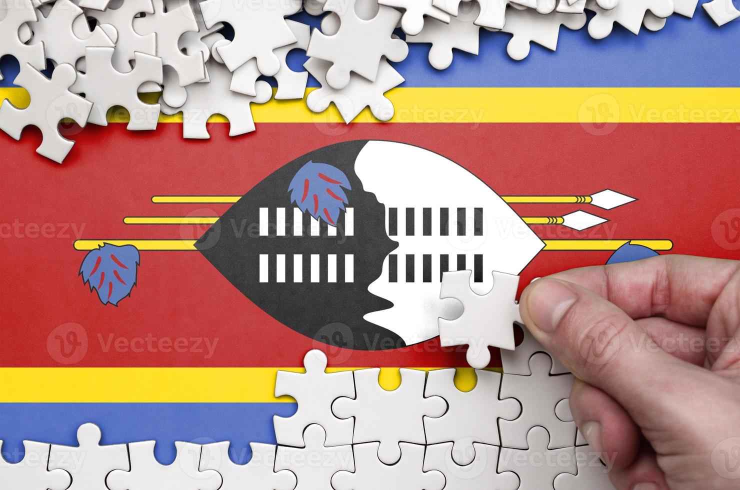 Swaziland flag  is depicted on a table on which the human hand folds a puzzle of white color photo