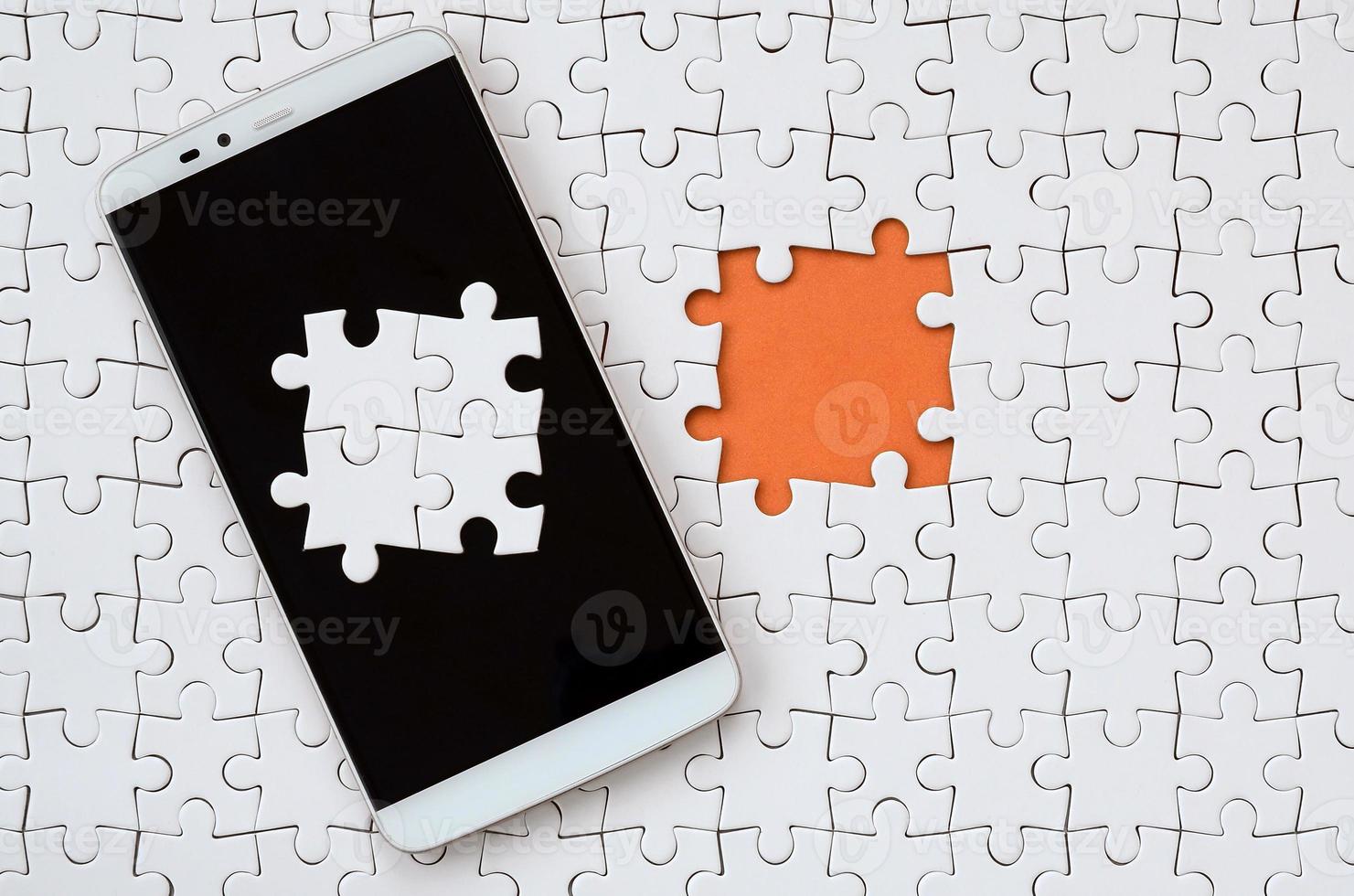 A modern big smartphone with several puzzle elements on the touch screen lies on a white jigsaw puzzle in an assembled state with missing elements photo