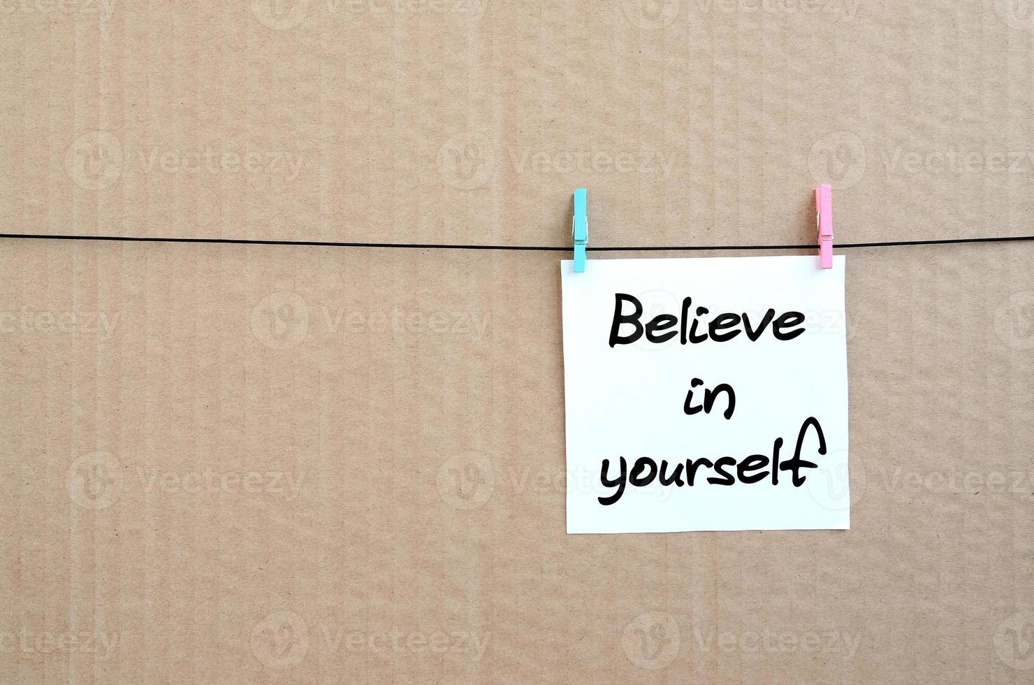 Believe in yourself. Note is written on a white sticker that hangs with a clothespin on a rope on a background of brown cardboard photo