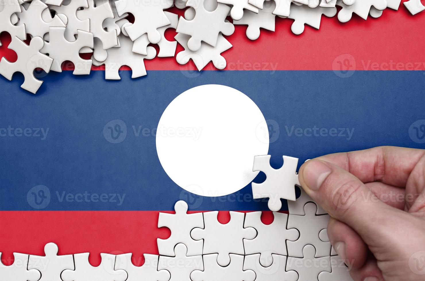 Laos flag  is depicted on a table on which the human hand folds a puzzle of white color photo