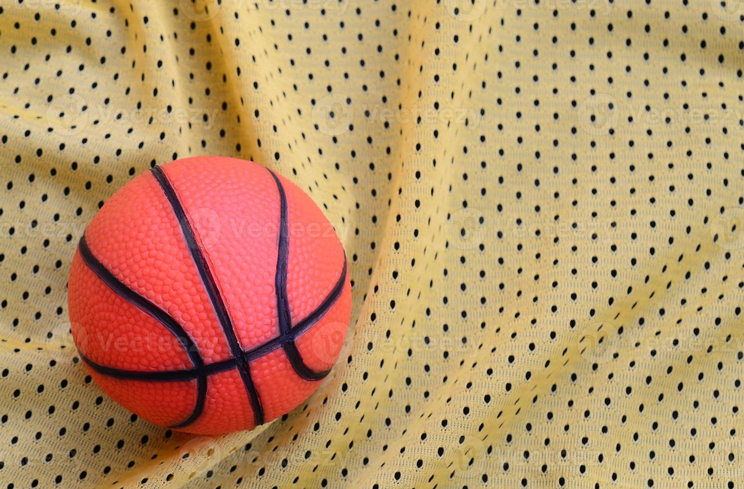 Small orange rubber basketball lies on a yellow sport jersey clothing fabric texture and background with many folds photo