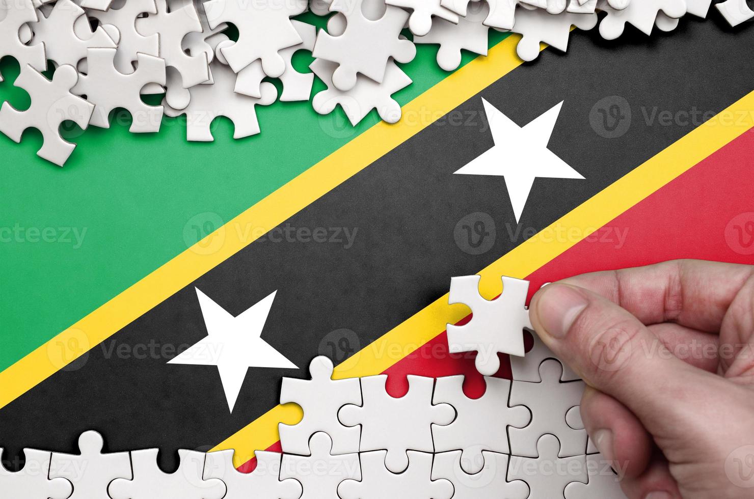Saint Kitts and Nevis flag  is depicted on a table on which the human hand folds a puzzle of white color photo