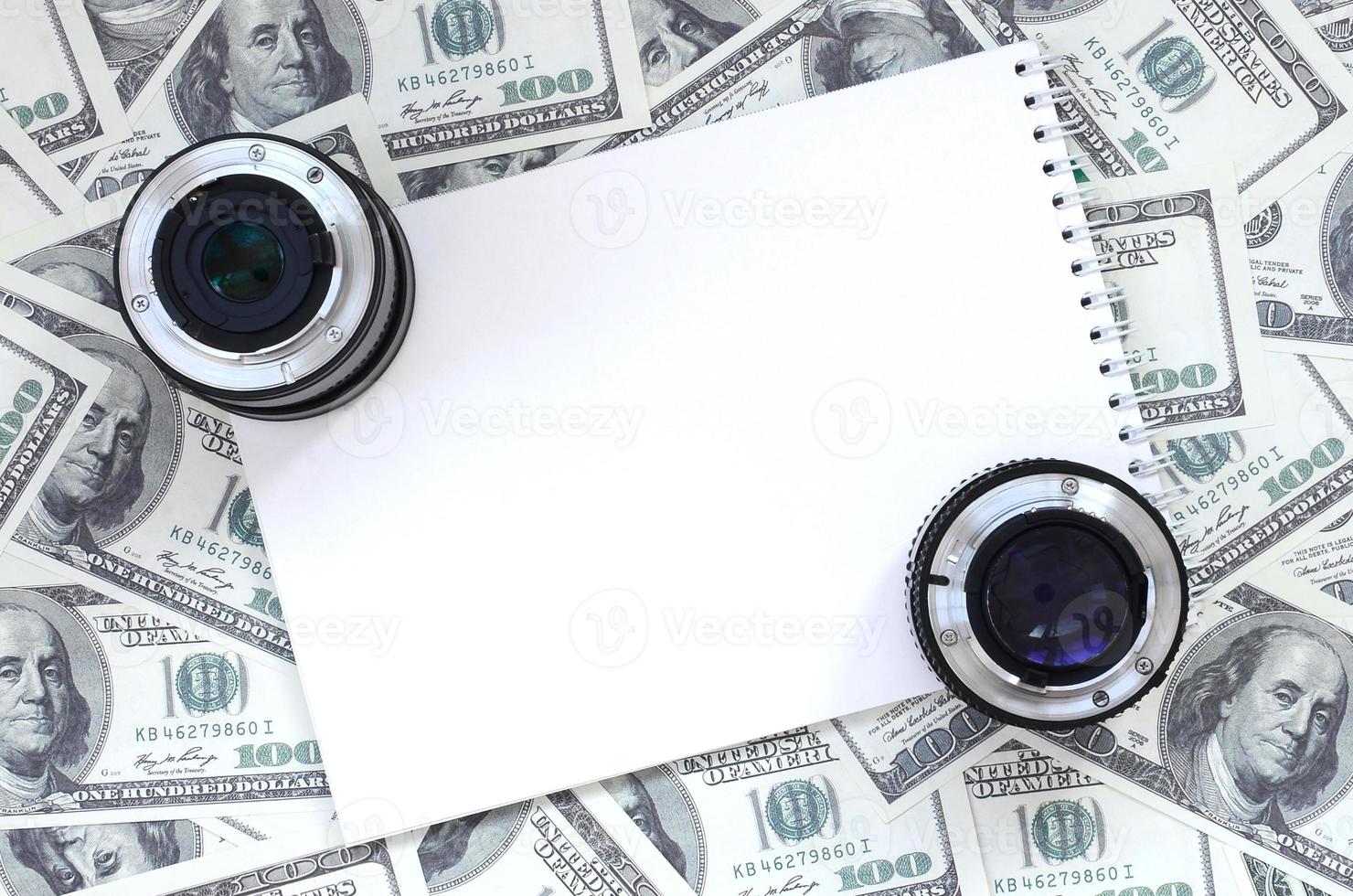 dos lentes fotográficos y un cuaderno blanco se encuentran en el fondo de muchos billetes de dólar. espacio para texto foto