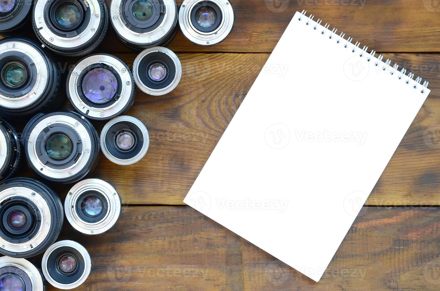 Several photographic lenses and white notebook lie on a brown wooden background. Space for text photo