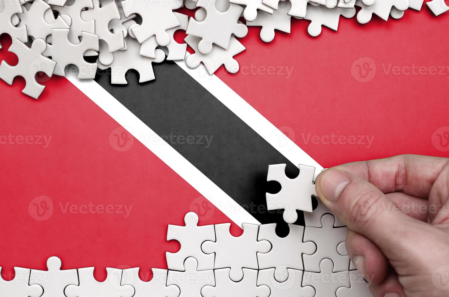 Trinidad and Tobago flag  is depicted on a table on which the human hand folds a puzzle of white color photo
