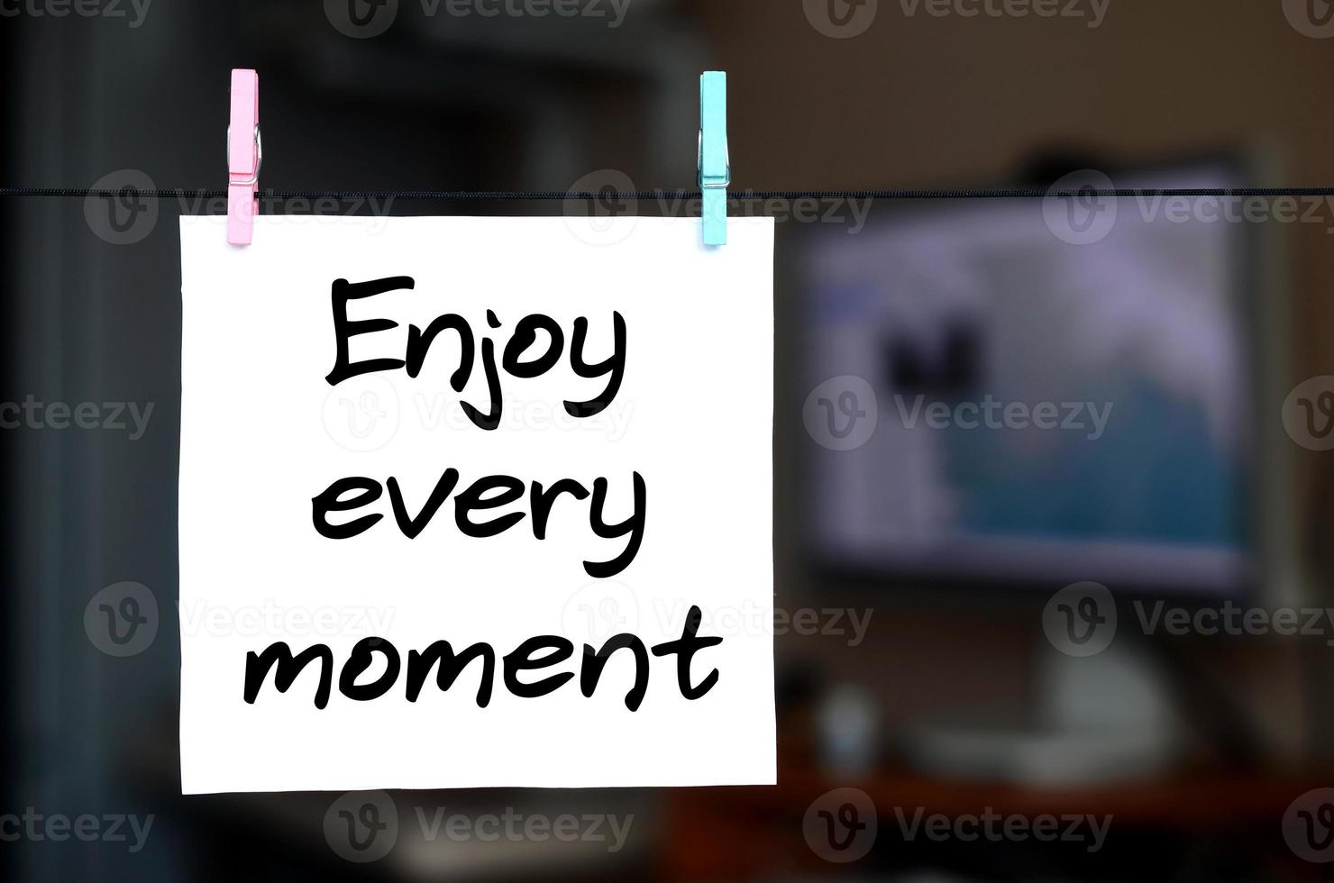 Enjoy every moment. Note is written on a white sticker that hangs with a clothespin on a rope on a background of office interior photo