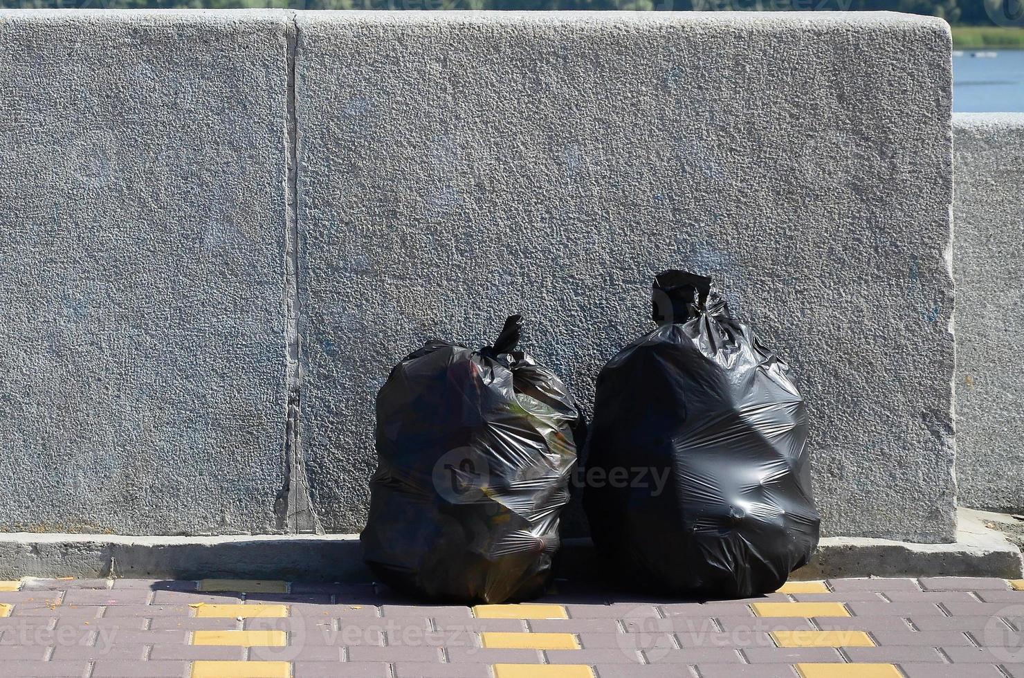 Two black garbage bags on tiled street floor at concrete fence in city photo