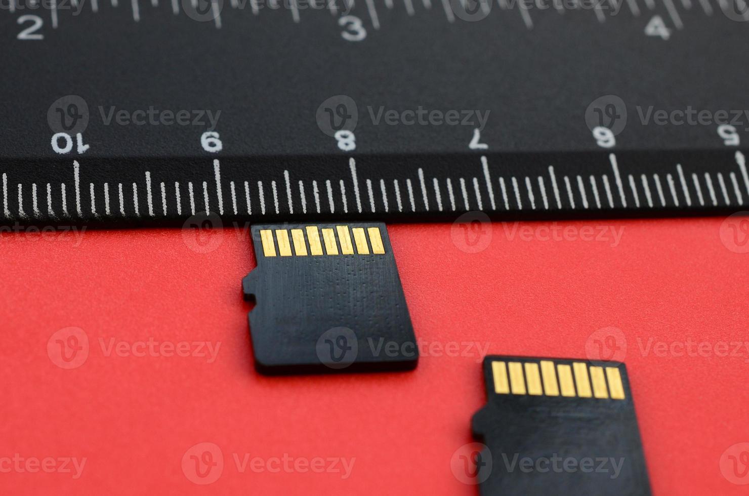 Two small micro SD memory cards lie on a red background next to a black ruler. A small and compact data and information store photo