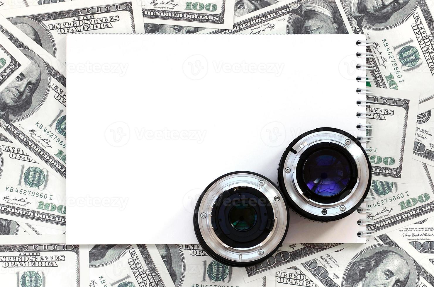 Two photographic lenses and white notebook lie on the background of a lot of dollar bills. Space for text photo