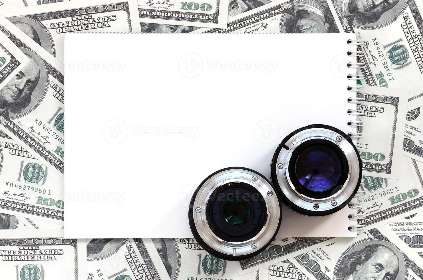 dos lentes fotográficos y un cuaderno blanco se encuentran en el fondo de muchos billetes de dólar. espacio para texto foto