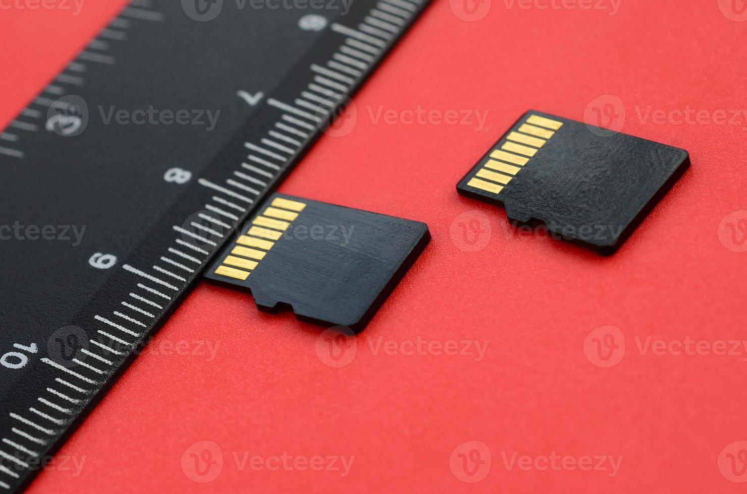 Two small micro SD memory cards lie on a red background next to a black ruler. A small and compact data and information store photo