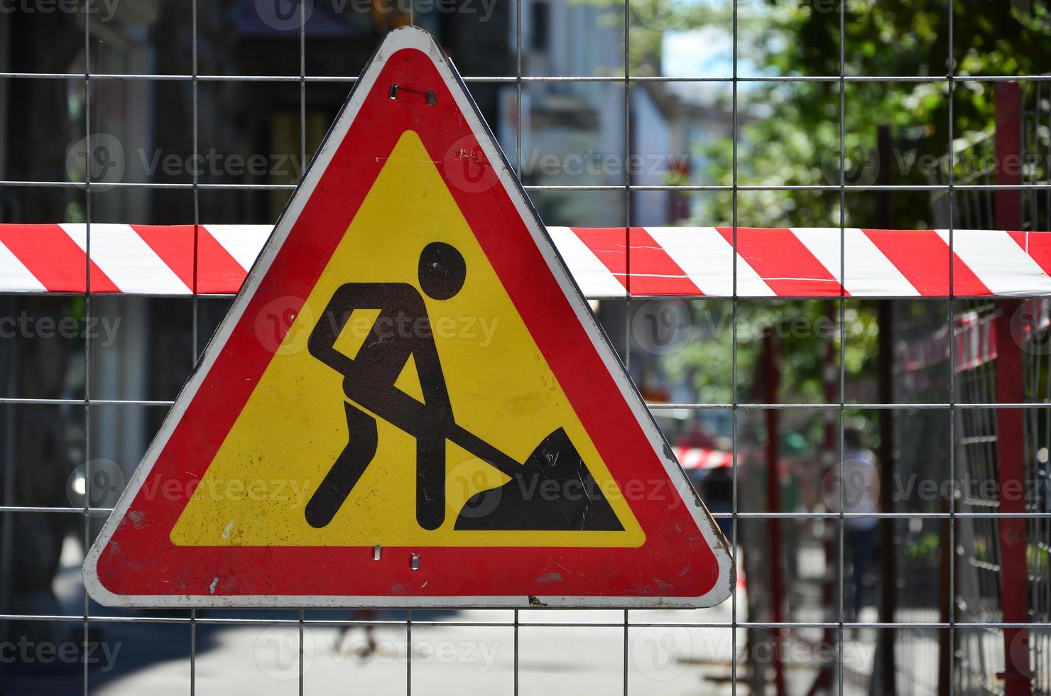 The warning sign under construction is attached to a metal mesh fence with a red and white striped signal tape photo