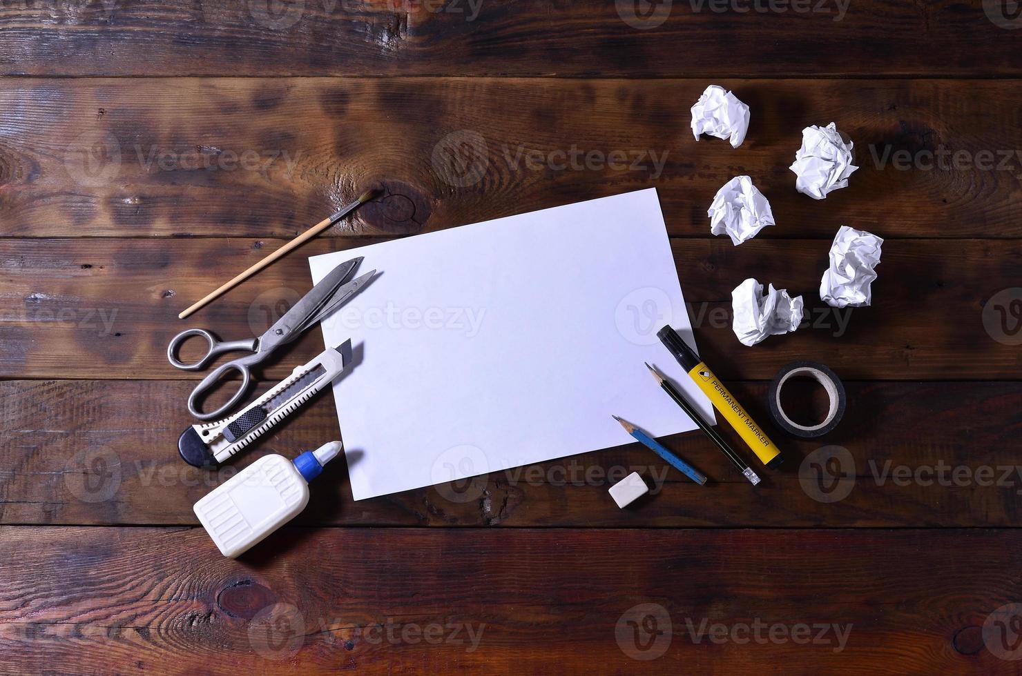 A school or office still life with a white blank sheet of paper and many office supplies. The school supplies lie on a brown wooden background. Place for text photo