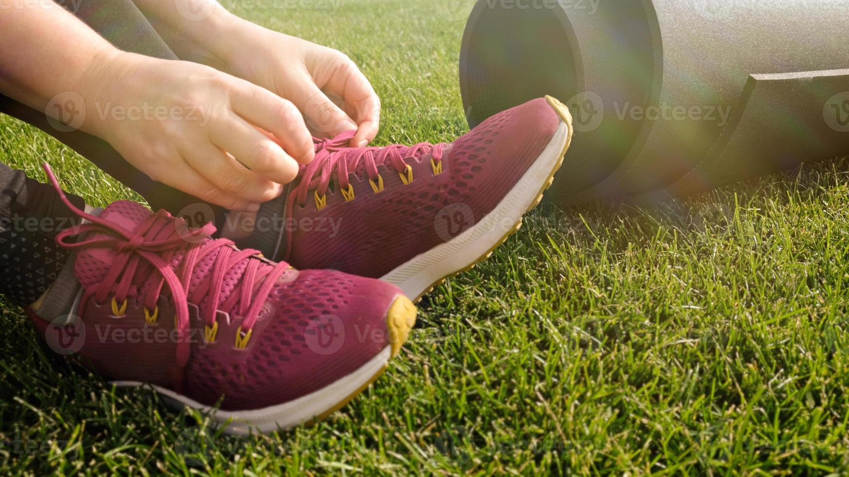 Young woman ties the laces on the pink sneaker. Woman preparing for yoga asana or fitness workout on green lawn in the backyard or park. Warm up before running. Healthy lifestyle. photo