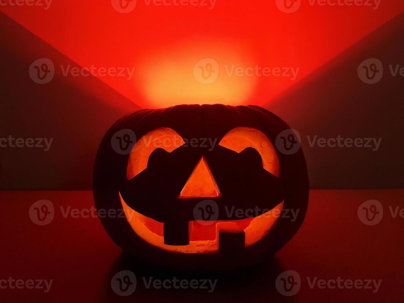 Halloween holiday background with orange smiling jack-o-lantern. Big carved glowing pumpkin stands centered on the surface in dark night. The back wall highlighted in red triangle over pumpkin. photo