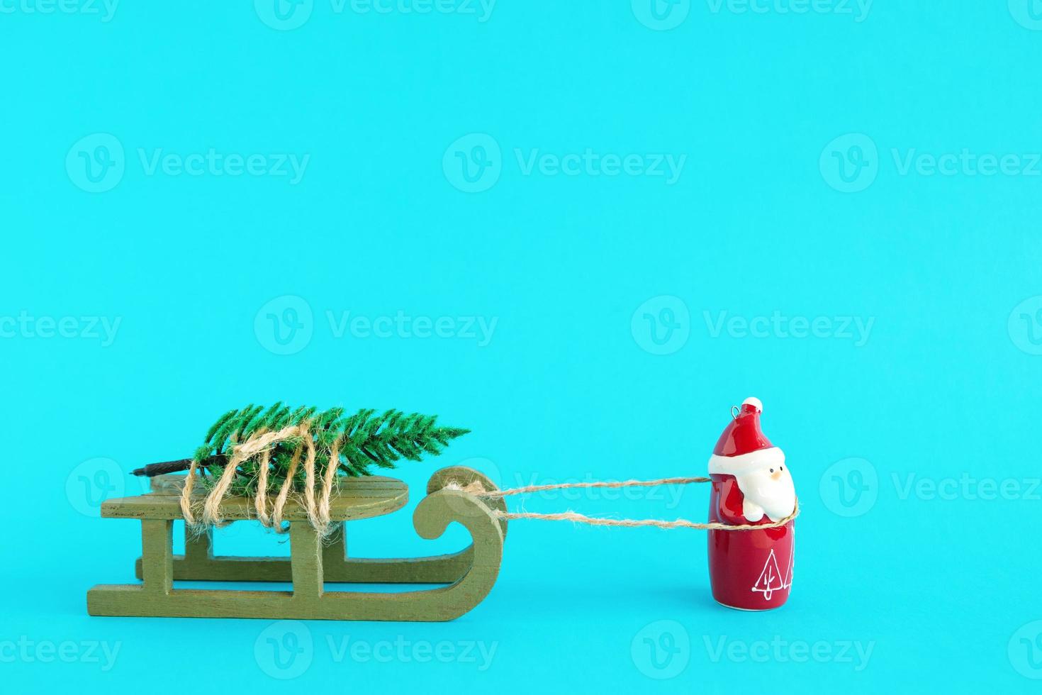 santa claus lleva un árbol de navidad en un trineo de madera verde sobre un fondo azul brillante. concepto de navidad y año nuevo con espacio de copia. foto