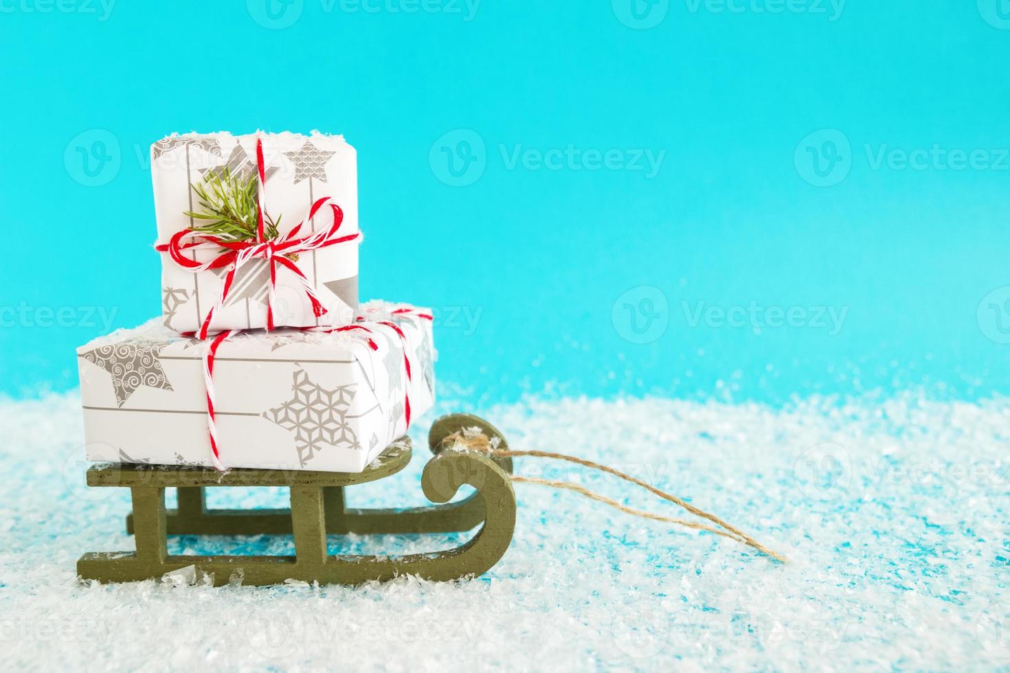 Two gift boxes wrapped of white and gray paper with red-white ribbon, decorated of fir branch on a green wooden sleigh on a blue background with artificial snow. Christmas and New Year concept. photo