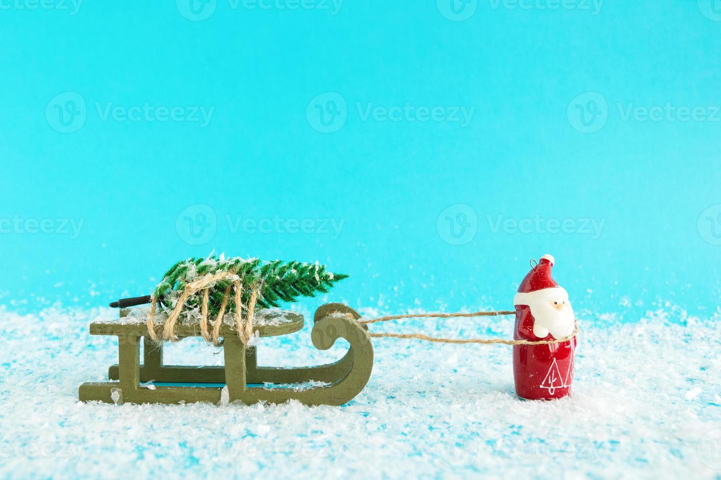Santa Claus is carrying a Christmas tree on a green wooden sleigh on a blue background with artificial snow. Christmas and New Year concept with copy space. photo