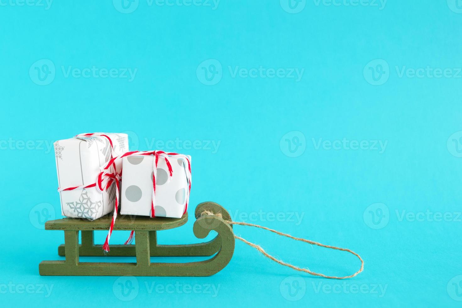 Two small gift boxes wrapped of white and gray paper with red-white ribbon on a green wooden sleigh on a bright blue background. Christmas and New Year concept with copy space. photo