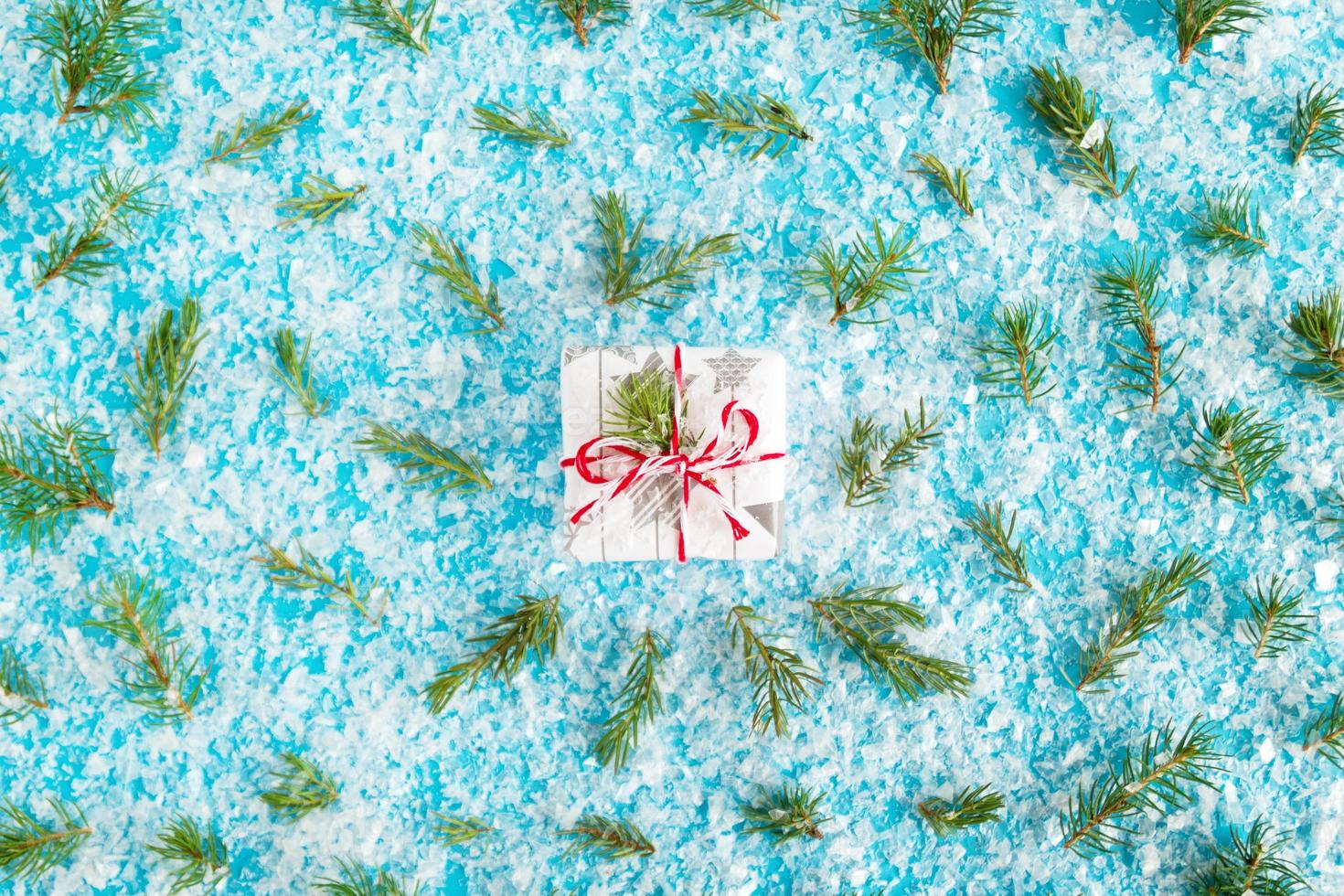 Small gift box wrapped of white and gray paper with red-white ribbon on the blue background with artificial snow decorated fir branches, top view. Christmas and New Year concept with copy space. photo