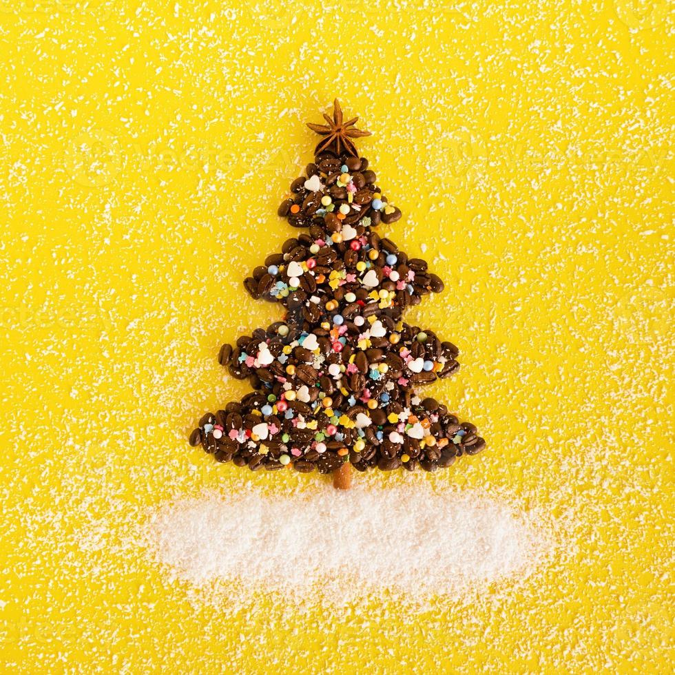 árbol de navidad hecho de granos de café y estrella de anís decorada, aspersión culinaria multicolor y chips de coco sobre un fondo amarillo, vista superior. foto