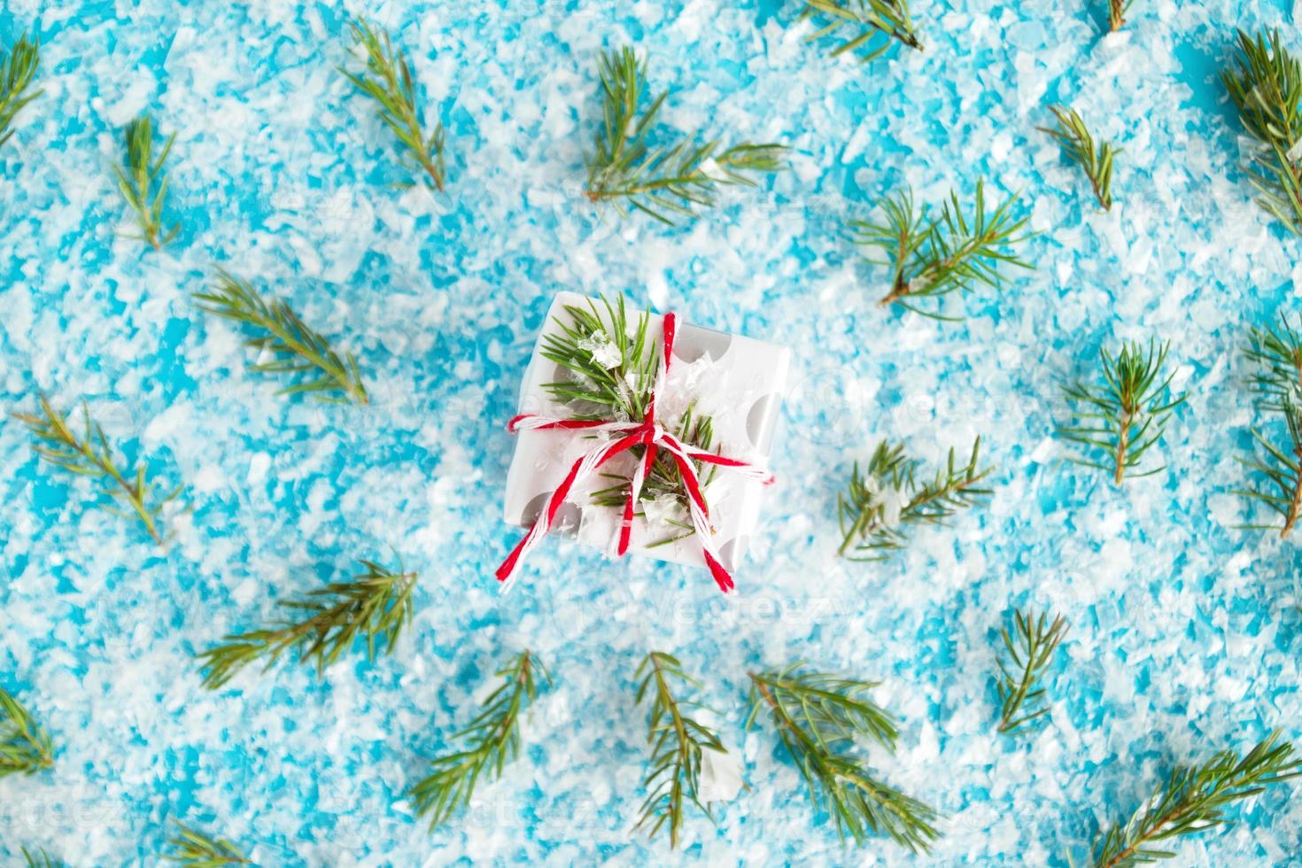 Small gift box wrapped of white and gray paper with red-white ribbon on the blue background with artificial snow decorated fir branches, top view. Christmas and New Year concept with copy space. photo