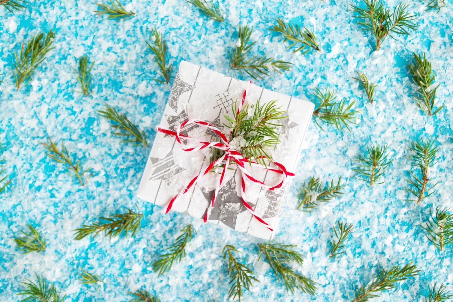 Gift box wrapped of white and gray paper with red-white ribbon on the blue background with artificial snow decorated fir branches, top view. Christmas and New Year concept with copy space. photo