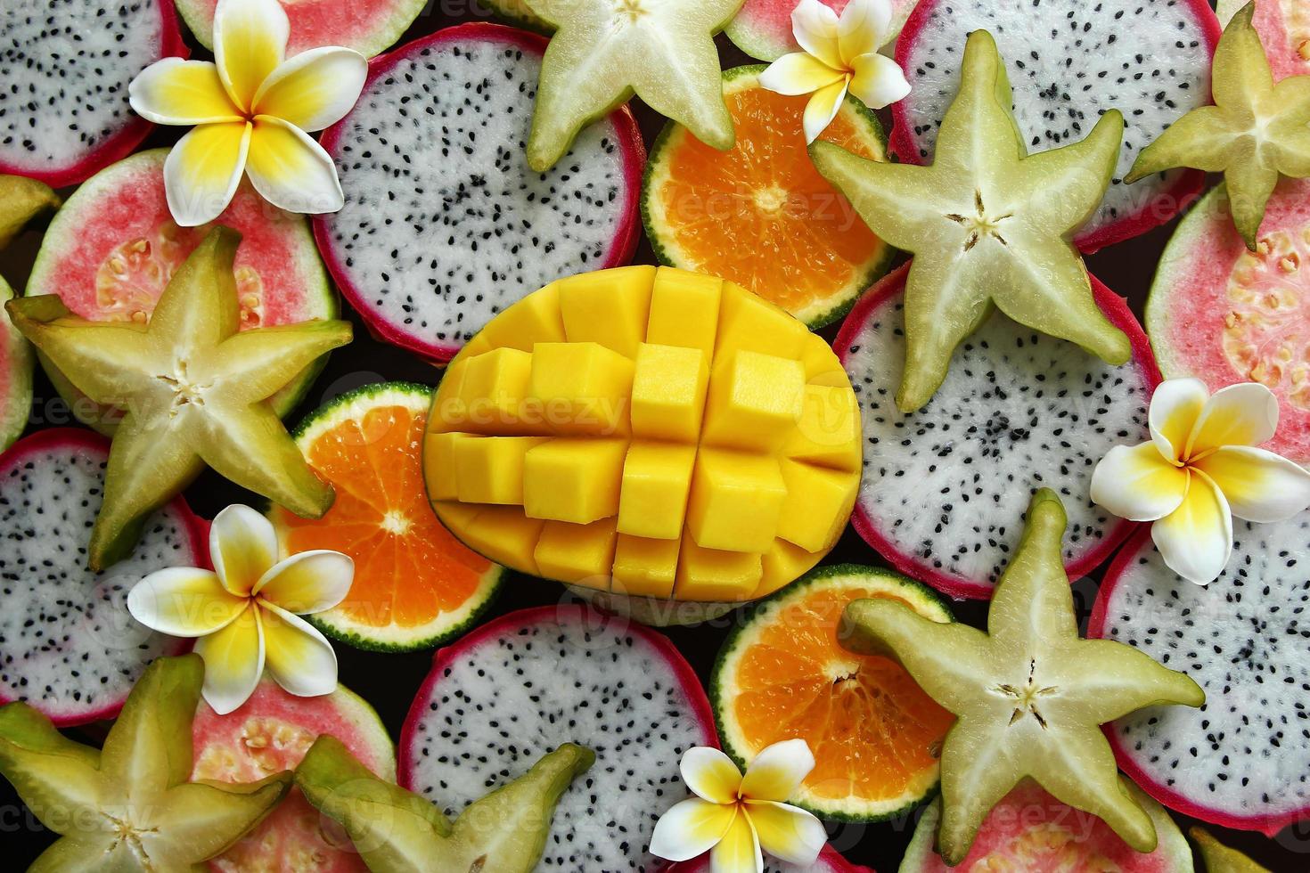 Top view on the mixed fresh and ripe tropical fruits - mango, tangerine, guava, dragon fruit, star fruit, sapodilla with flowers of plumeria. photo